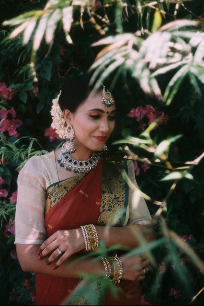 A South Asian woman in a red saree adorned with traditional jewelry amidst lush greenery.