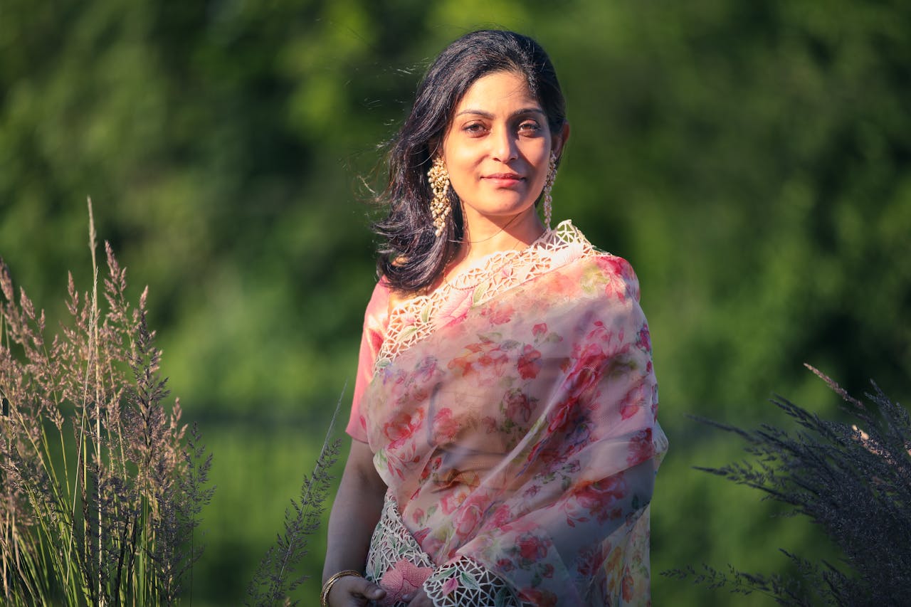 A woman in a floral saree standing amidst green foliage under sunlight.